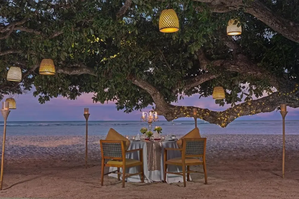 A beachside table set under a tree at Samabe Bali Suites & Villas, a Bali resort all-inclusive.