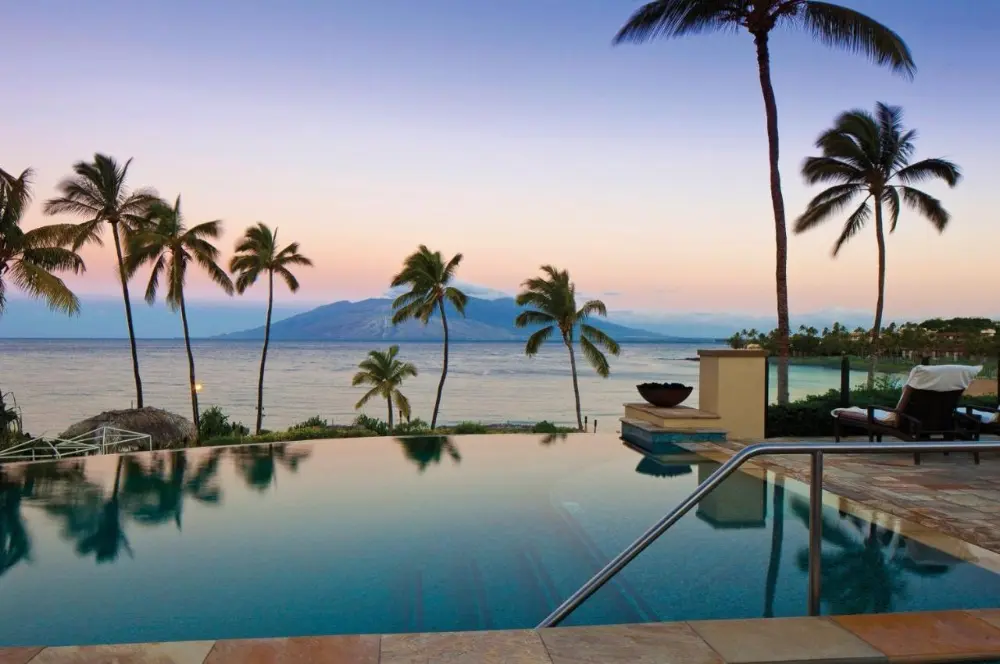 A serene sunset view of the pool and lounge chairs surrounded by palm trees at Four Seasons Resort Maui at Wailea, one of the best Four Seasons hotels in the world.