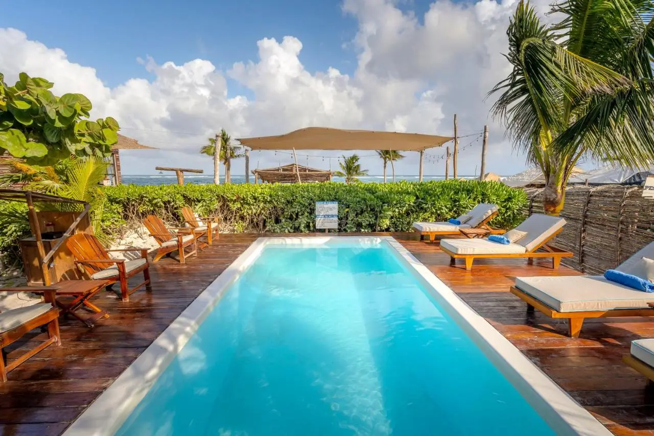 A sunny beach scene with a pool, lounge chairs, and umbrellas at Villa Pescadores, one of the best party hotels in Tulum.