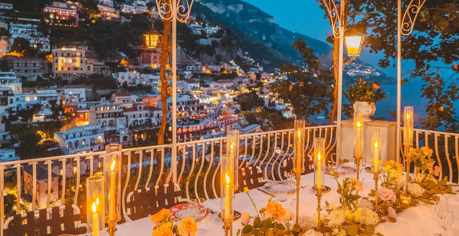 A candlelit table set for dinner at the Hotel Poseidon, one of the best romantic hotels in Positano, overlooks a hillside town with numerous lit buildings at dusk.