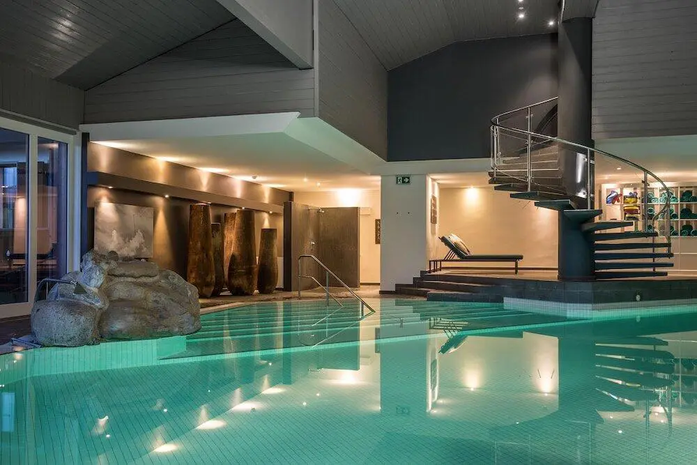 A beautiful indoor swimming pool with a spiral staircase, perfect for relaxing after a day at Huus Gstaad, one of the best snowboard hotels in Switzerland.