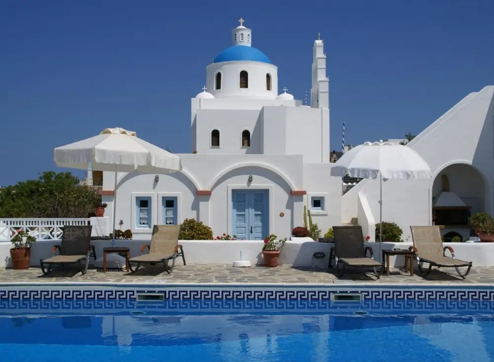 Aethrio Sunset Village, a family-friendly hotel in Santorini, featuring a blue and white building situated near a pool with umbrellas.