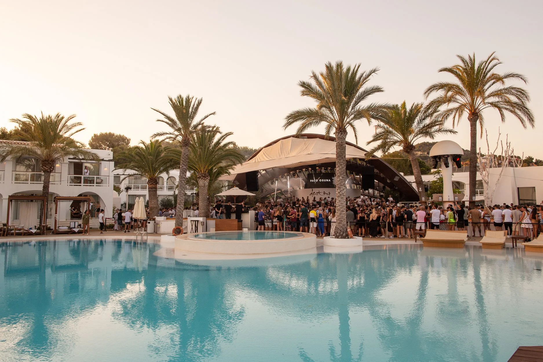 A lively pool area at Destino Pacha, with palm trees and people enjoying the festive ambiance under the sun in this party hotel in Ibiza