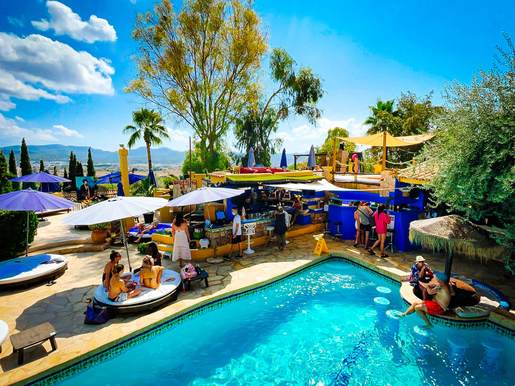 A vibrant pool scene at Pikes, Party Hotel in Ibiza, featuring guests relaxing on lounge chairs under colorful umbrellas.