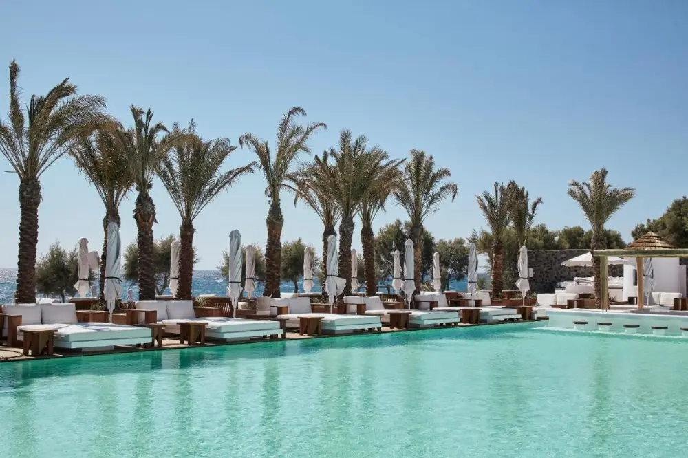 A stunning pool at Nikki Beach Resort, a spa hotel in Santorini, surrounded by lounge chairs and palm trees.