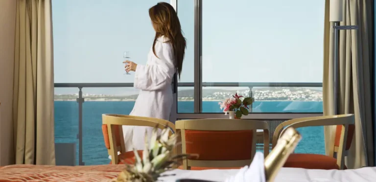 A woman in a white robe stands by a hotel room balcony, enjoying a glass of wine with an ocean view—an ideal way to increase hotel bookings by showcasing an exclusive guest experience.