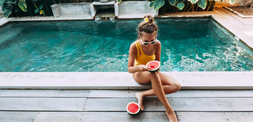 A young woman in a yellow swimsuit enjoys fresh watermelon by a private pool, capturing user-generated content that increases hotel bookings by inspiring future travellers with an authentic guest experience.